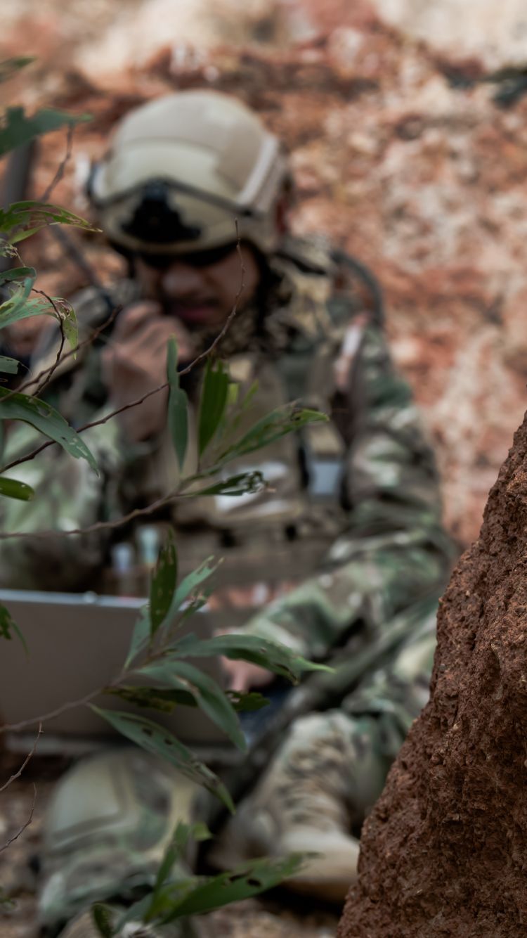 a soldier hidden between rocks and bushes using a rugged laptop